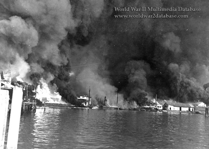 Fires at Cavite Navy Yard resulting from the Japanese air raid on December 10, 1941. Fifty-four bombers of the 11th Air Fleet were detailed from Formosa and attacked at 1300 hours. Twenty-seven attacked ships and small craft in the bay and the remainder went on toward Cavite, dropping their bombs from a height of 20,000 feet, above the range of the nine 3-inch anti-aircraft guns protecting the base. Almost every bomb fell within the Navy Yard. After the first run, the first flight of twenty-seven withdrew and the other twenty-seven bombers, having completed their attack against ships in the bay, flew in to strike the targets. The attack lasted for two hours. The entire yard was set ablaze; the power plant, dispensary, repair ships, warehouses, barracks, and radio station received direct hits. Greatest damage was done by the fire which spread rapidly and was soon out of control. Admiral Rockwell estimated that five hundred men were killed or seriously wounded. The covered self-propelled lighter YF-181 - perhaps visible in the right center - is loaded with almost 200 burning torpedos, which will be consumed in the flames, crippling the offensive capability of the United States Navy Asiatic Fleet's submarines. At the time this photograph was taken, small arms ammunition was exploding in the center of the heavy blaze on the left. The submarine whose bow is visible at the far right is probably USS Sealion (SS-195), hit by bombs and had settled by the stern. Sealion, a 1450-ton Sargo class submarine, was commissioned in late November 1939 and, in the spring of 1940, deployed to the Far East to strengthen the defenses of the Philippines as relations with Japan deteriorated. Sealion was nested at Machina Wharf with USS Seadragon (SS-194) inboard and minesweeper USS Bittern (AM-36) outboard. Most Sealion personnel were below decks. The first stick of bombs landed from 100 to 200 yards (90 to 180 meters) astern of Sealion; all hands were ordered all hands below. On the second